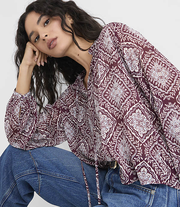 A woman in a patterned blouse and jeans rests her head on her hand against a plain background.