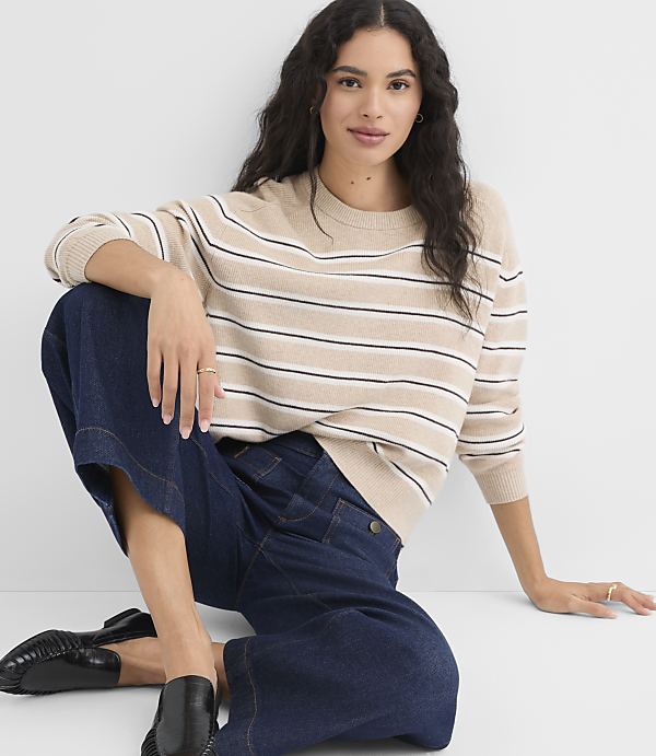 A woman in a striped sweater and wide-leg jeans sits on a white floor against a plain white background.