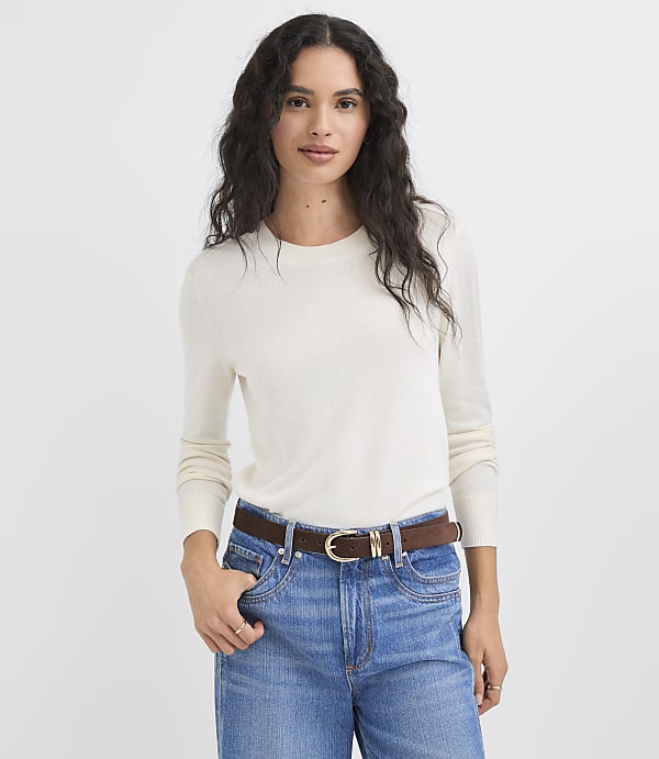 A woman in a cream sweater and blue jeans stands against a plain white background.