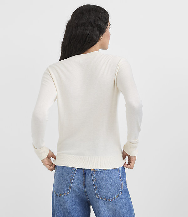 A woman in a cream-colored sweater and blue jeans stands against a plain white background.