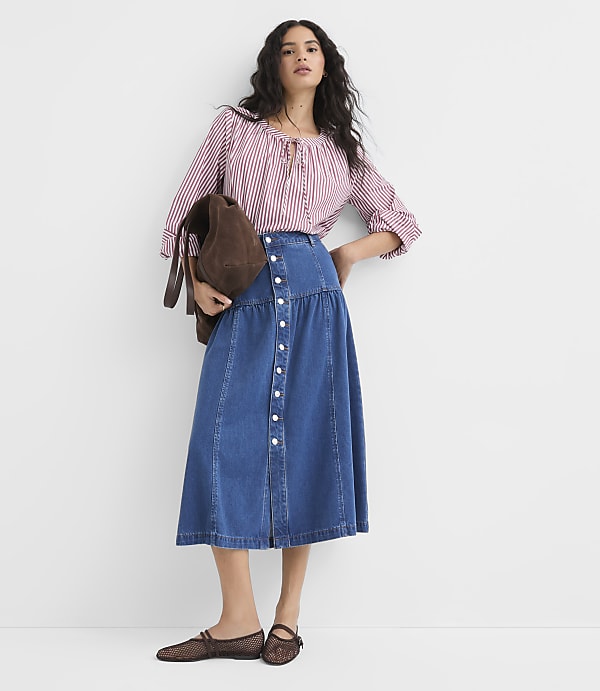 A woman in a striped blouse and denim skirt stands against a plain white background, holding a brown bag.