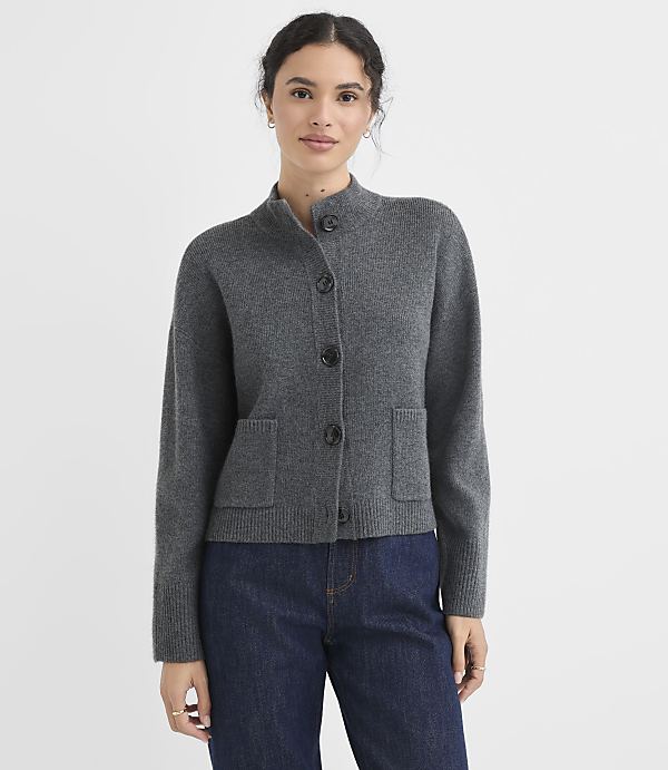 A woman in a gray cardigan and blue jeans stands against a plain white background.
