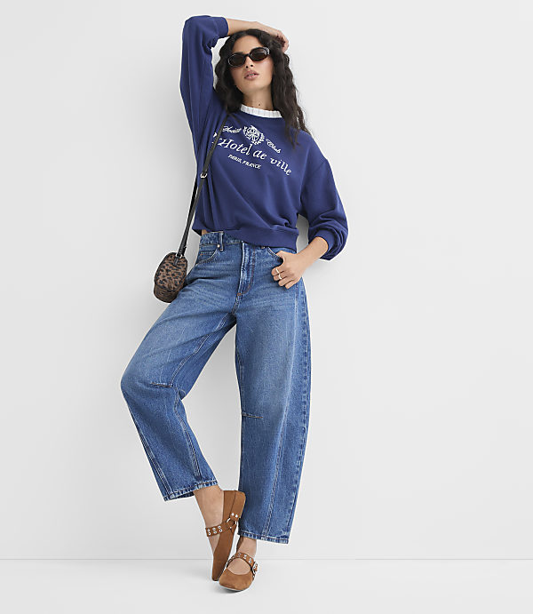 A woman in a blue sweatshirt and wide-leg jeans poses against a plain white background.