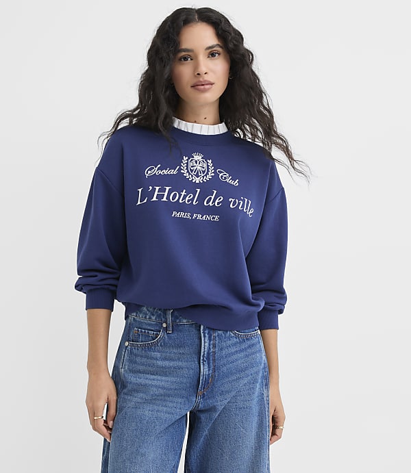 A woman in a blue sweatshirt and jeans stands against a plain white background.