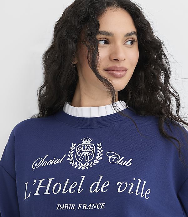 A woman in a blue sweatshirt with a white ruffled collar stands against a plain white background.