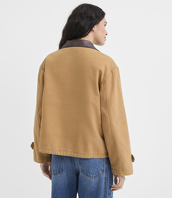 A woman in a tan jacket and blue jeans stands against a plain white background.