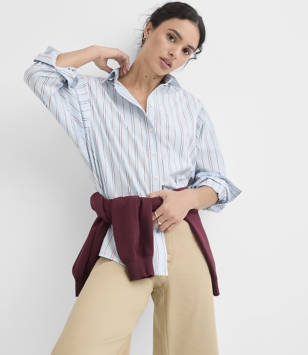 A woman in a light blue pinstripe shirt and tan pants poses against a plain white background, with a maroon sweater tied around her waist.