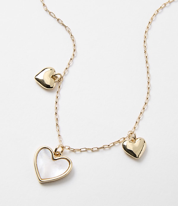 A delicate gold chain necklace with three heart-shaped charms, one inlaid with mother-of-pearl, rests against a plain white background.