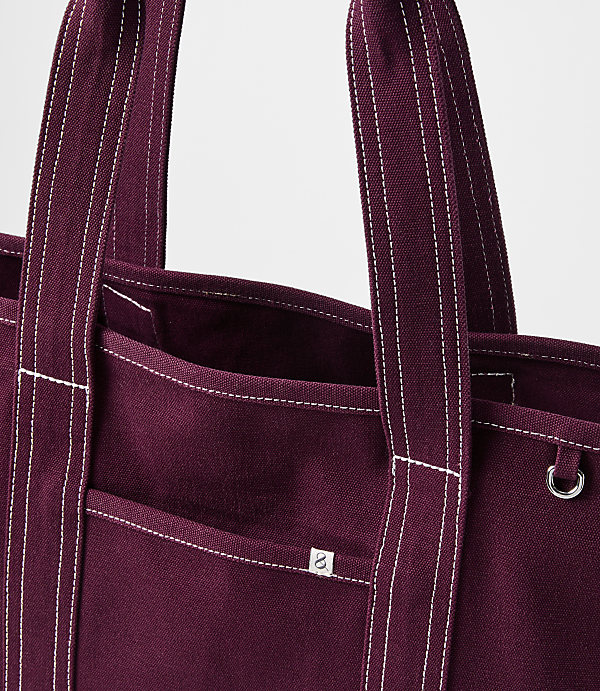 A close-up of a burgundy canvas tote bag with white stitching and a small logo is presented against a plain white background.