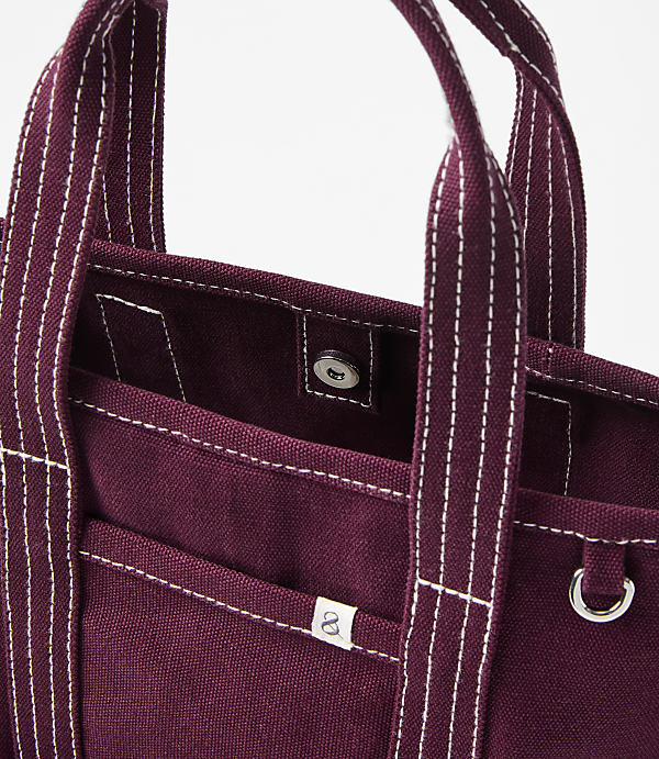 A close-up of a burgundy canvas tote bag with white stitching and a silver snap closure is set against a plain white background.