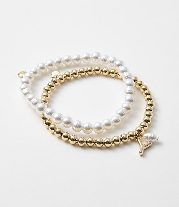 Two bracelets, one of pearls and one of gold beads with a charm, rest on a plain white background.