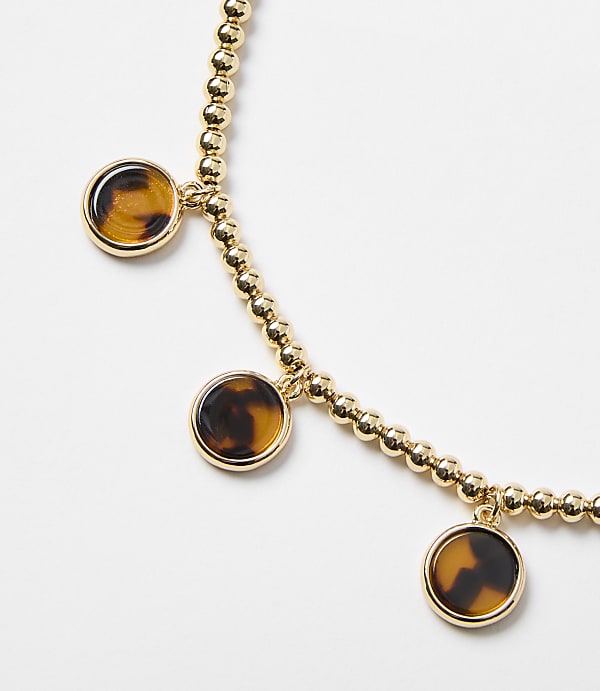 A gold beaded bracelet with three tortoise shell charms hangs against a plain white background.