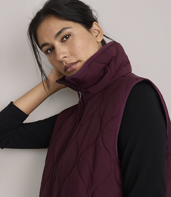 A woman in a burgundy quilted vest and black top poses against a plain, light background.