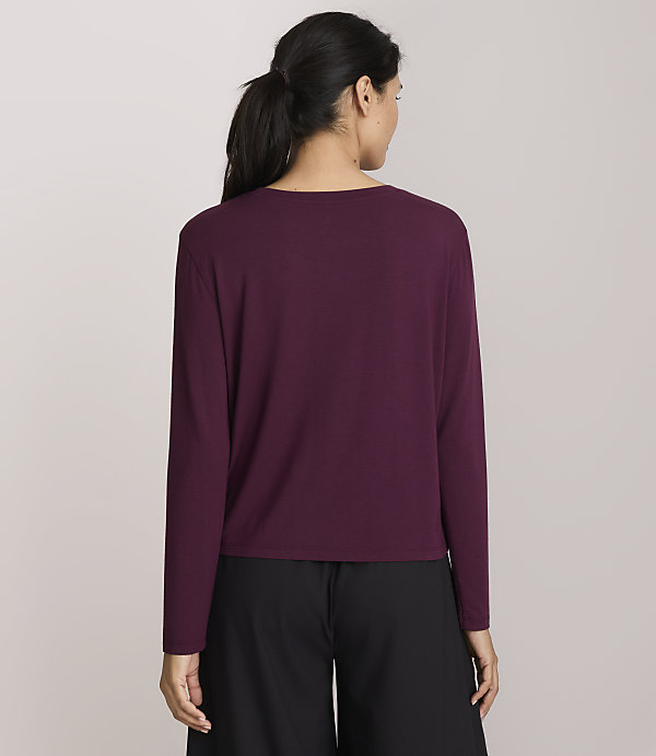 A woman in a maroon long-sleeved shirt and black pants stands against a plain, light purple background.