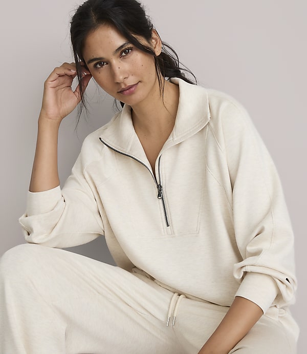 A woman in a cream-colored quarter-zip sweatshirt and matching pants poses against a plain, light gray background.
