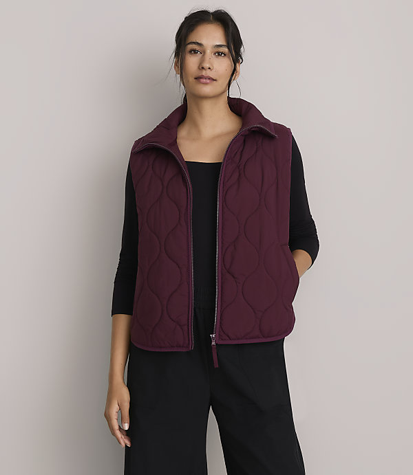 A woman in a quilted vest and hoodie stands against a plain purple background, holding a striped sweater.