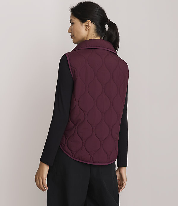 A woman in a quilted burgundy vest and black clothing stands against a plain, light-colored background.