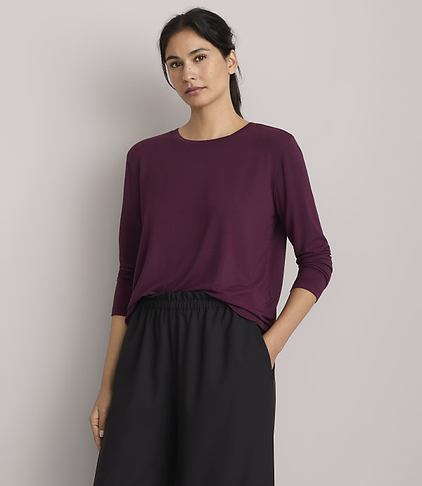 A woman in a plum-colored shirt and black pants stands against a plain gray background.