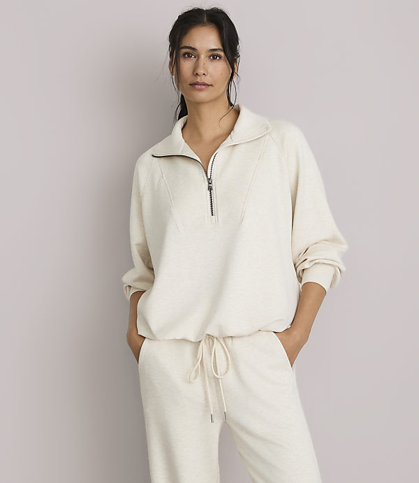 A woman in a cream-colored quarter-zip sweatshirt and matching pants stands against a plain, light gray background.
