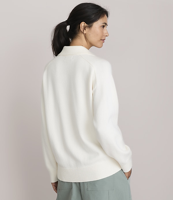 A woman in a cream sweater and light green pants stands against a plain, light pink background.
