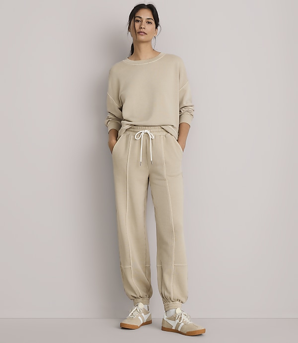 A woman in a beige sweatshirt and sweatpants stands against a plain gray background.