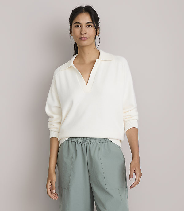 A woman in a cream-colored collared sweater and green pants stands against a plain, light gray background.