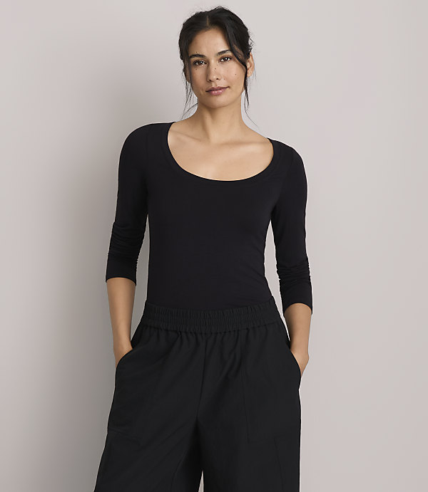 A woman in a black scoop-neck shirt and black pants stands against a plain gray background.