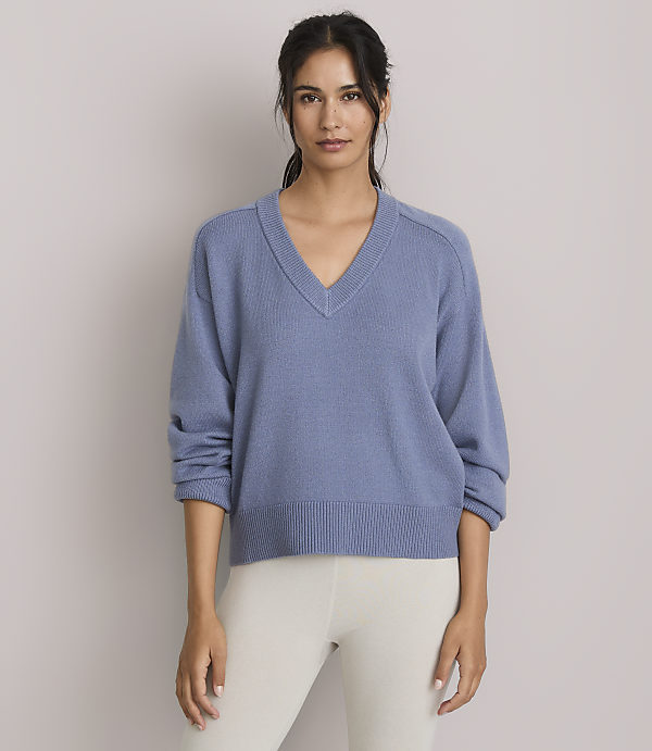 A woman in a blue v-neck sweater and cream leggings stands against a plain gray background.