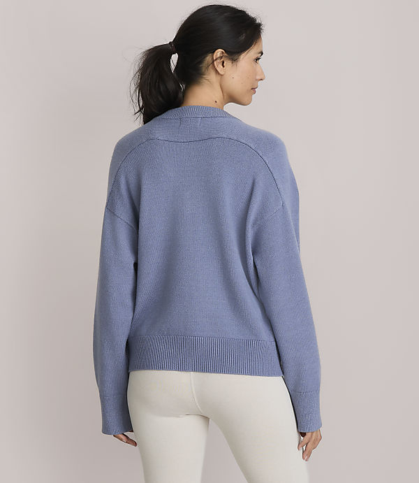 A woman in a blue sweater and white leggings stands against a plain, light pink background.