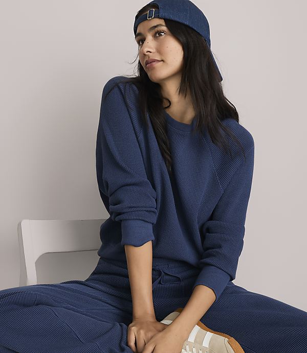 A woman in a blue waffle-knit sweatshirt and matching pants, wearing a blue baseball cap, sits on a white chair against a plain white background.