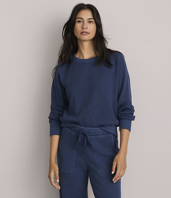 A woman in a blue sweatshirt and matching pants stands against a plain gray background.