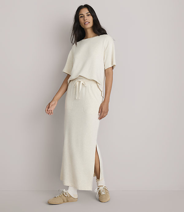 A woman in a cream-colored t-shirt and matching maxi skirt with a side slit stands against a plain, light gray background.