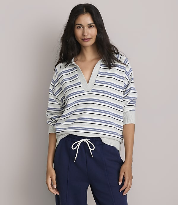 A woman in a striped collared shirt and navy sweatpants stands against a plain gray background.