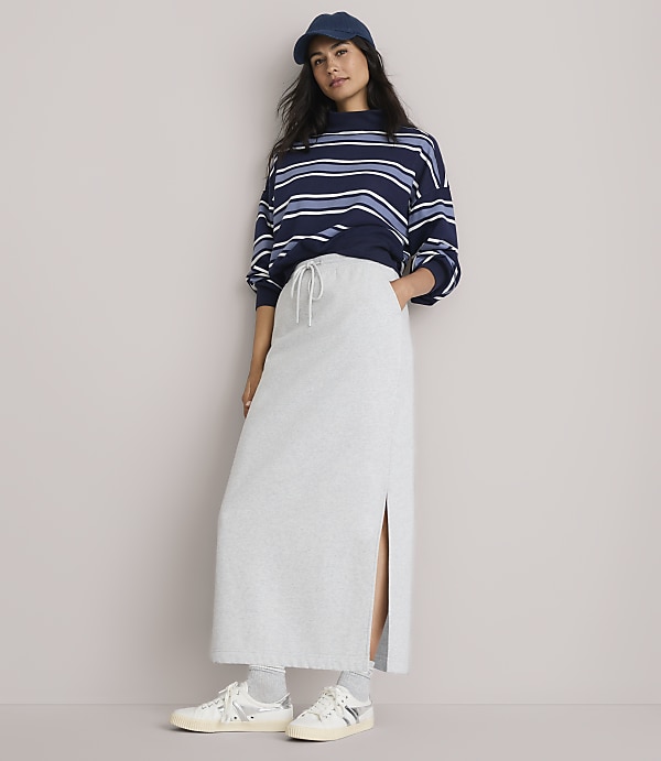 A woman in a striped sweatshirt and a light gray maxi skirt with a side slit stands against a plain, light gray background.