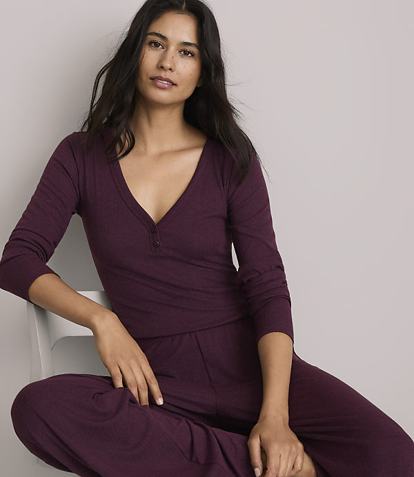 A woman in a burgundy henley and matching pants sits against a plain, light gray background.