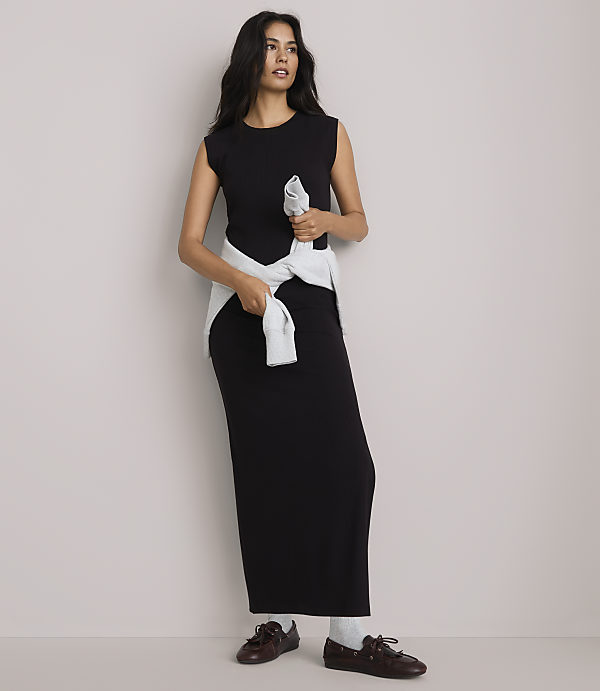 A woman in a black maxi dress and light blue sweater tied around her waist stands against a plain, light gray background.