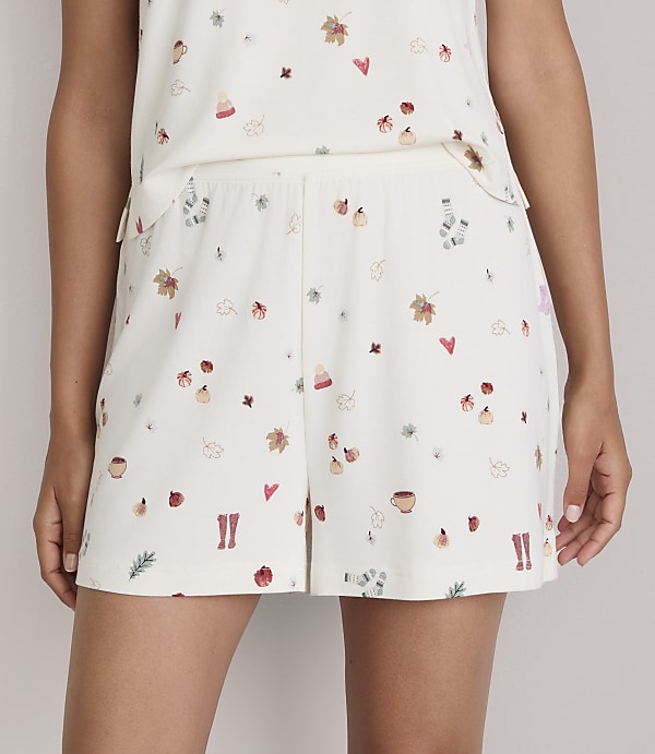 A person wears cream-colored shorts and a matching top adorned with a pattern of autumn leaves, hearts, boots, and cups, against a plain background.