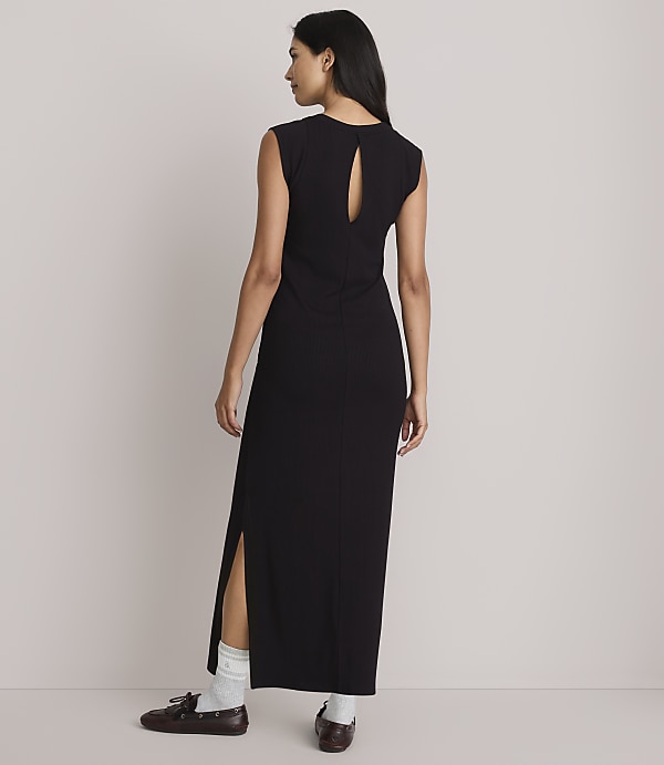 A woman in a black maxi dress with a cutout back and side slits stands with her back to the camera, wearing white socks and brown loafers against a plain, light-colored background.