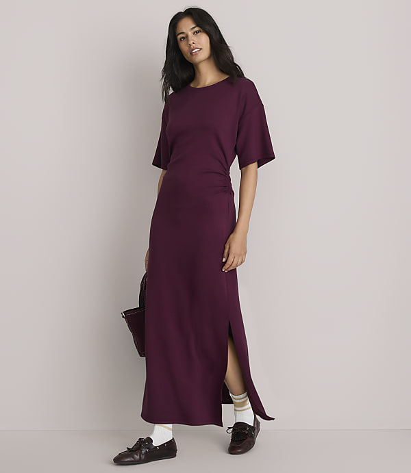 A woman in a plum-colored maxi dress with a side slit stands against a plain, light gray background, holding a dark bag.