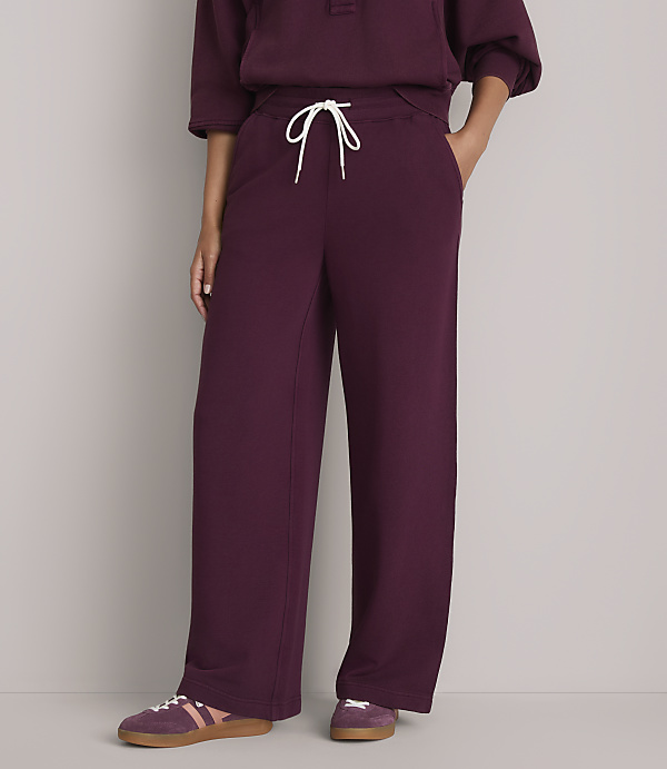 A person wearing burgundy sweatpants with a white drawstring stands against a plain gray background, with their feet in burgundy sneakers.