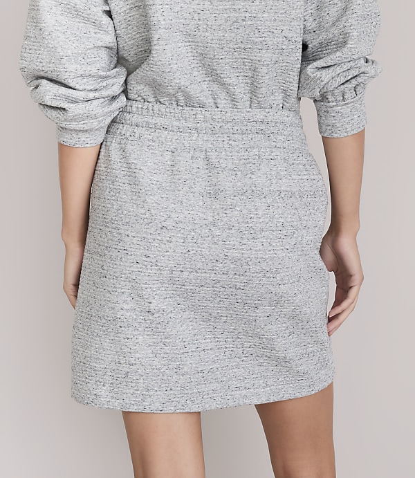 A person stands against a plain gray background, wearing a gray textured dress with puffed sleeves.