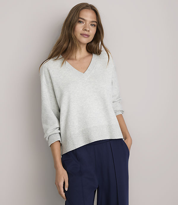 A woman in a light gray v-neck sweater and navy blue pants stands against a plain white background.