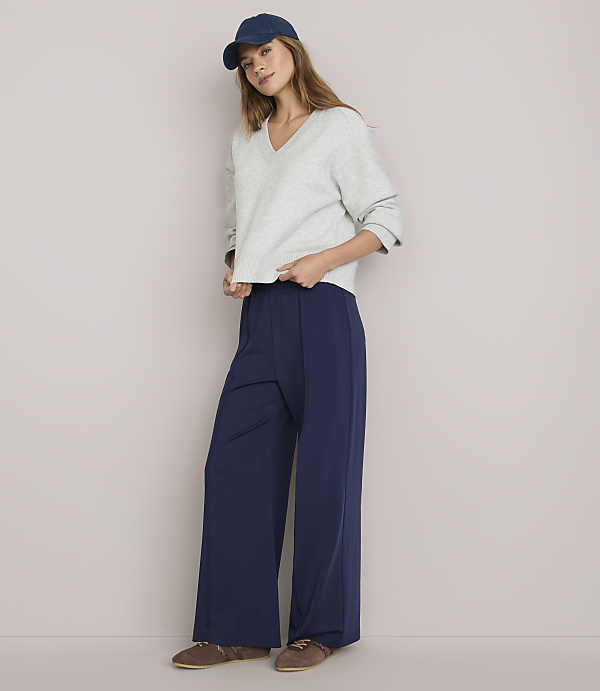 A young woman in a light gray sweatshirt, navy blue wide-leg pants, and a blue baseball cap stands against a plain white background.
