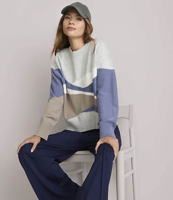A woman in a patterned sweater and navy pants sits on a white chair against a plain white background.