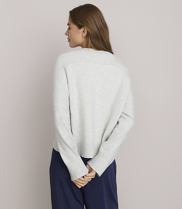 A woman in a light gray sweater and navy pants stands with her back to the camera against a plain white background.