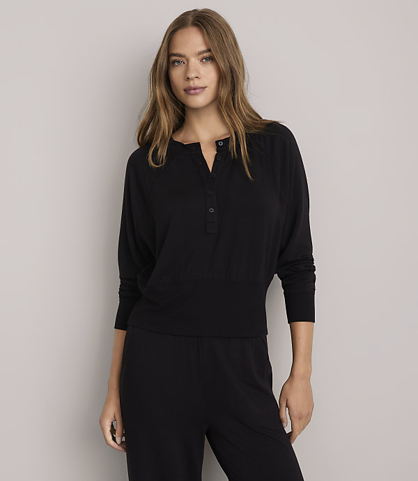 A woman in a black henley shirt and pants stands against a plain white background.