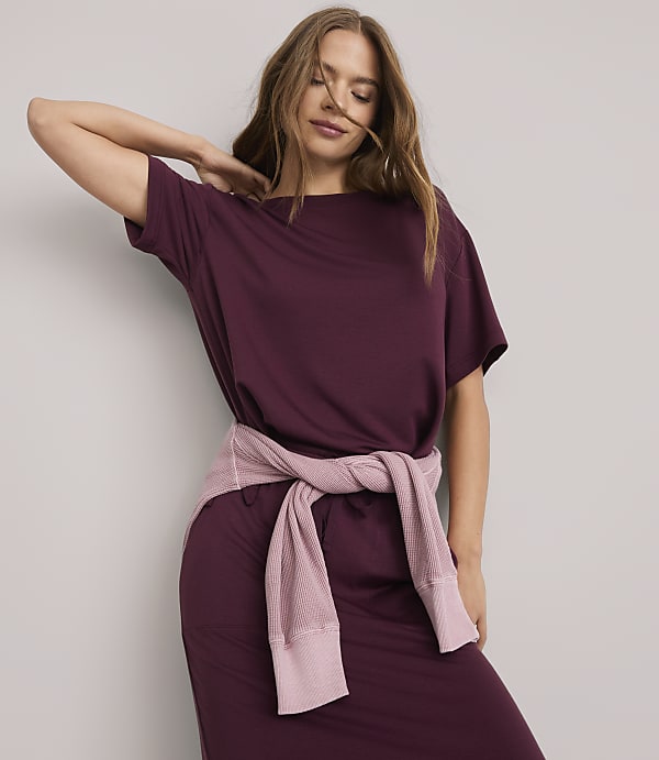 A woman in a burgundy dress with a pink sweater tied around her waist poses against a plain white background.