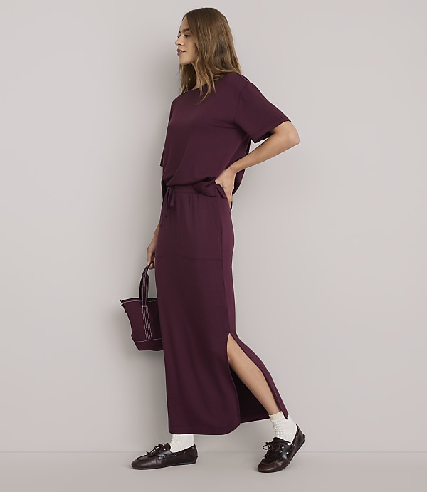 A woman in a burgundy t-shirt and matching maxi skirt with a side slit stands against a plain white background, holding a small burgundy bag.