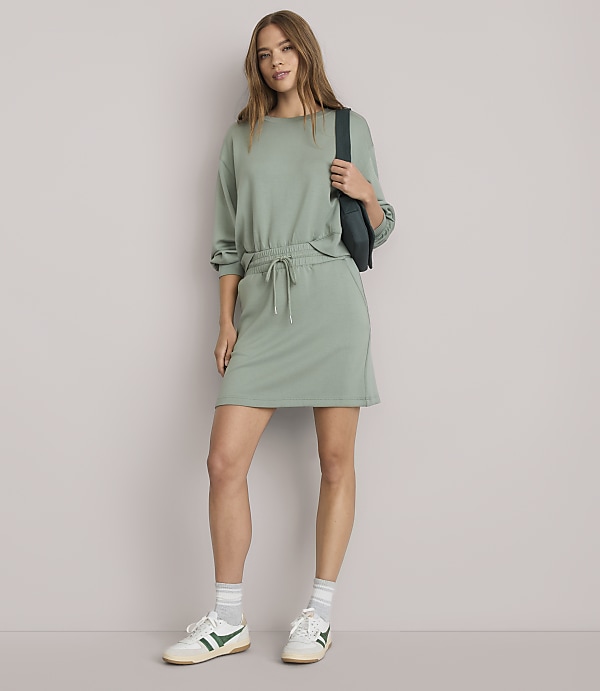 A woman in a light green sweatshirt dress and sneakers stands against a plain white background, with a dark green fanny pack slung across her chest.