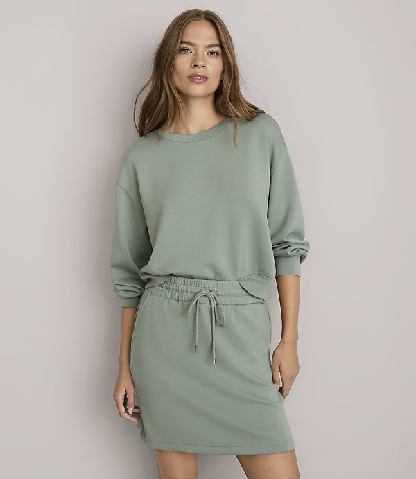 A woman in a green sweatshirt and skirt stands against a plain white background.